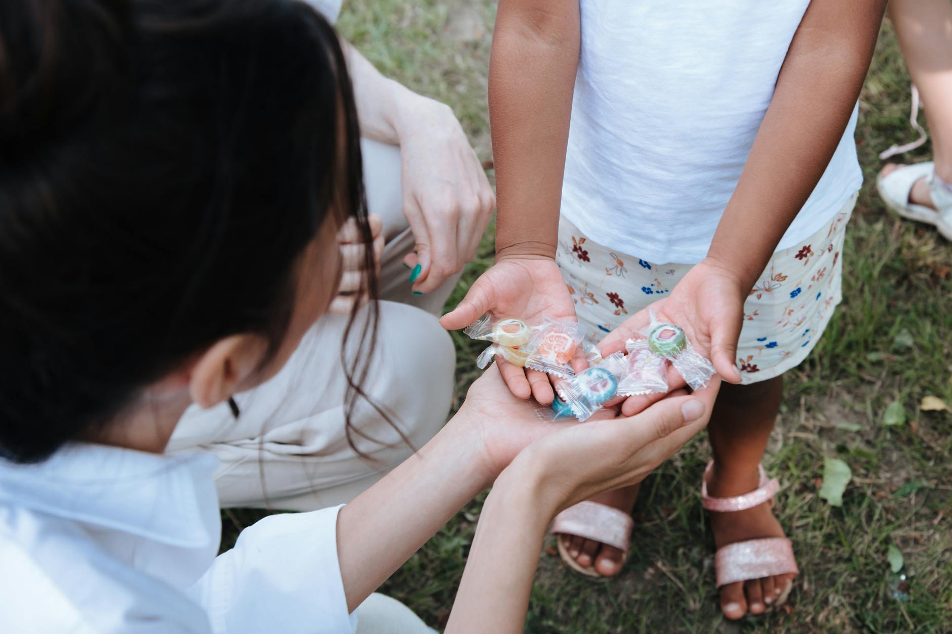 A child shares colorful wrapped candies with an adult in a park setting.