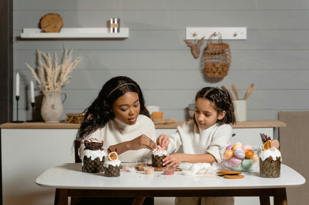 An African American woman and a Hispanic girl creatively decorate cupcakes in a cozy indoor setting.