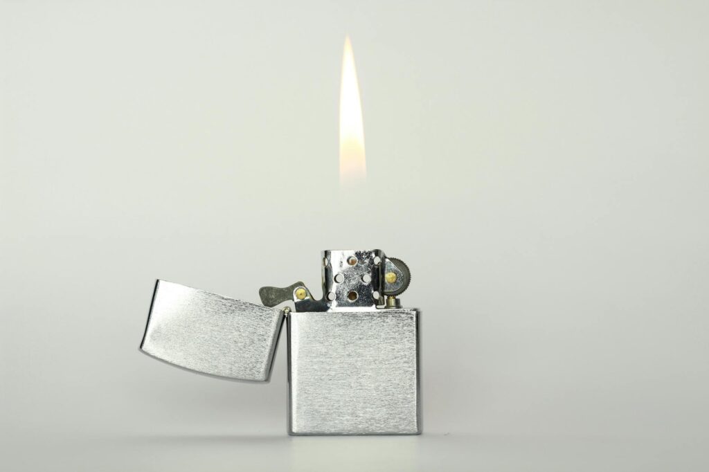 Close-up of a silver flip top lighter with a visible flame, isolated on a minimalistic background.