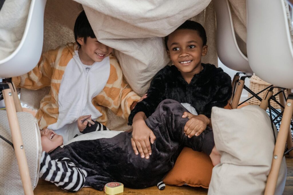 Three kids having fun inside a makeshift indoor fort with pillows, enjoying playful moments.