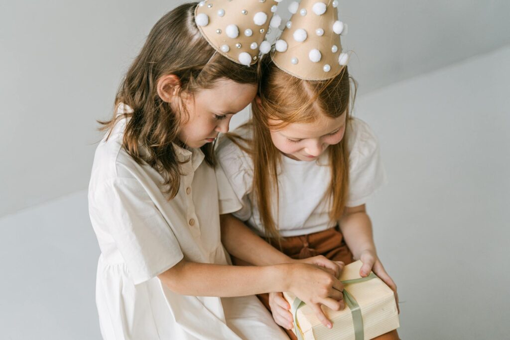 Two young girls in party hats joyfully opening a gift, embodying birthday celebration cheer.