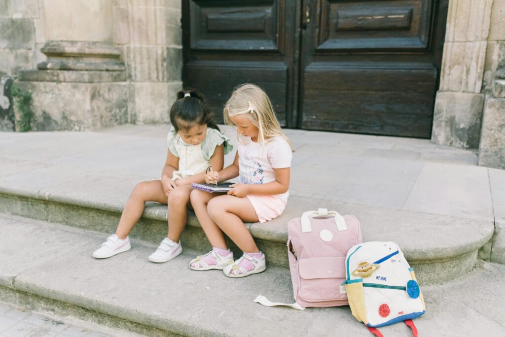 Two young girls sitting outside on steps, sharing a tablet with backpacks nearby.