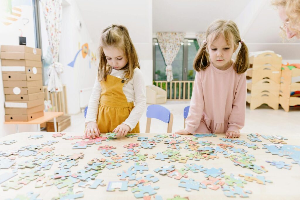 Two young girls solving puzzles at a kindergarten table with focus and creativity.