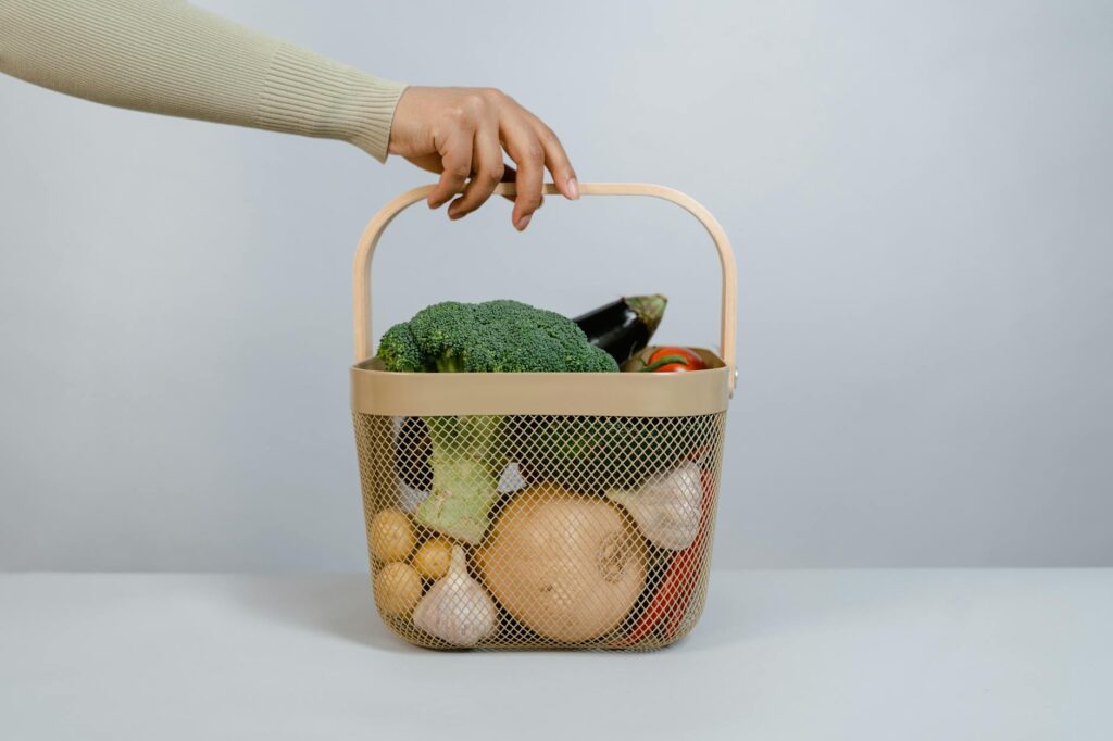 A hand holding a basket filled with fresh vegetables like broccoli and onions.