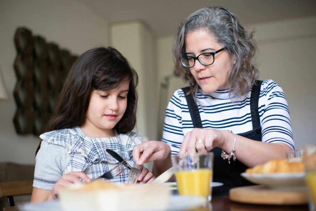 Grandmother guides young girl in table manners during breakfast at home.