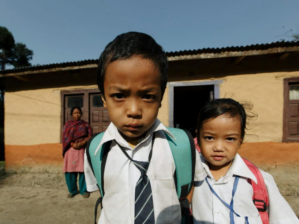 Two children wearing school uniforms stand outside a rural house, ready for school.