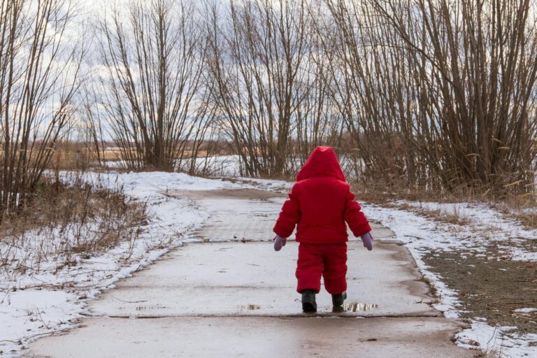 A child in a red outfit walks on a snowy path in a winter landscape with bare trees.