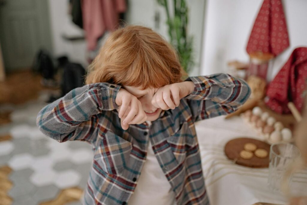 A young boy with red hair wipes his eyes in a cozy kitchen, evoking emotion and childhood innocence.