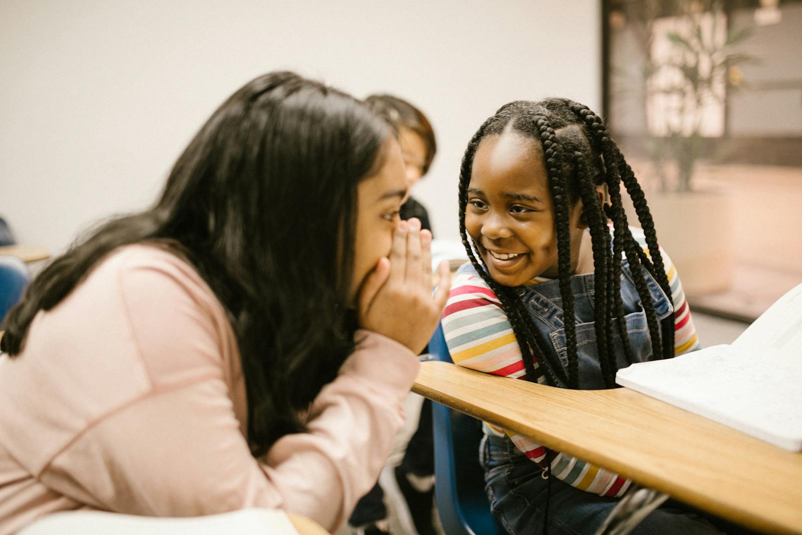 Two girls whisper and smile in a classroom, fostering friendship and joy.