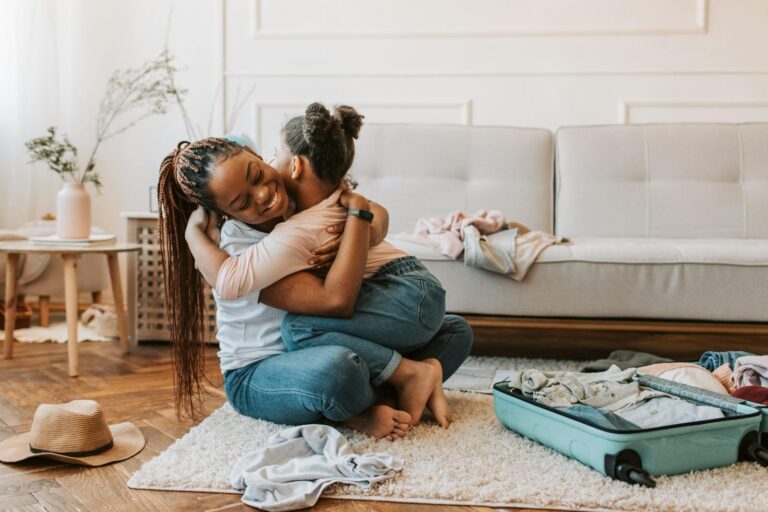 A mother and daughter share a loving hug while preparing for a trip with an open suitcase at home.