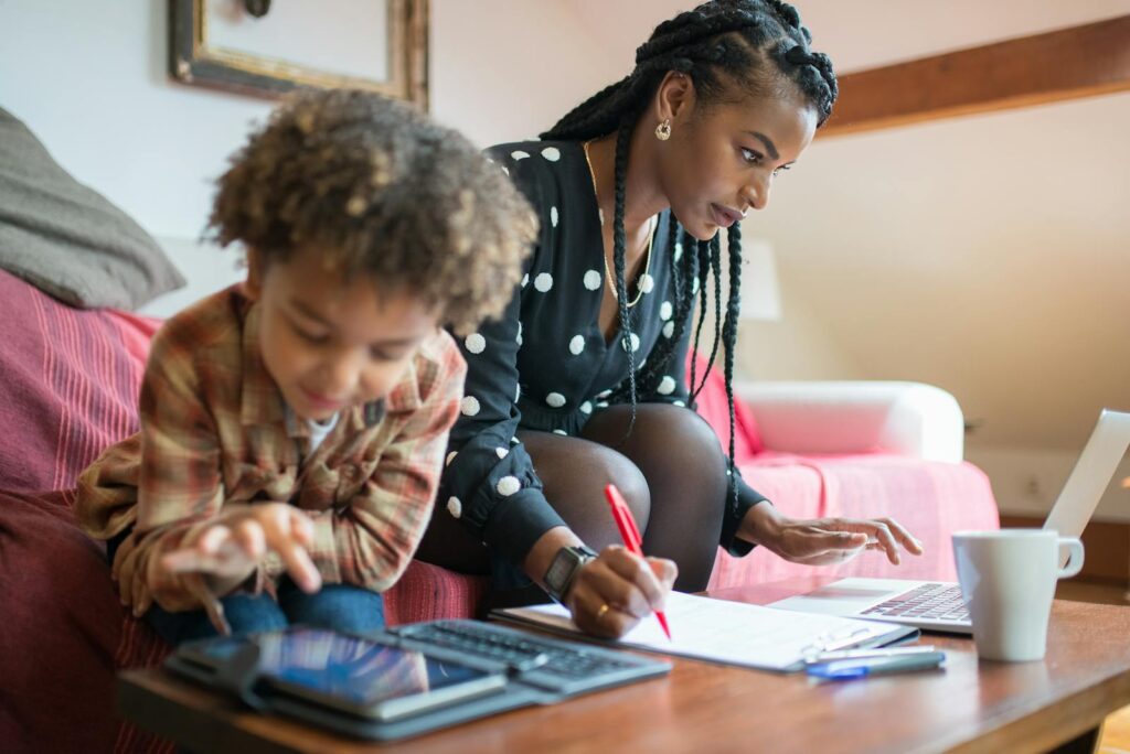 A mother multitasking at home, working on a laptop while her child plays on a tablet nearby.