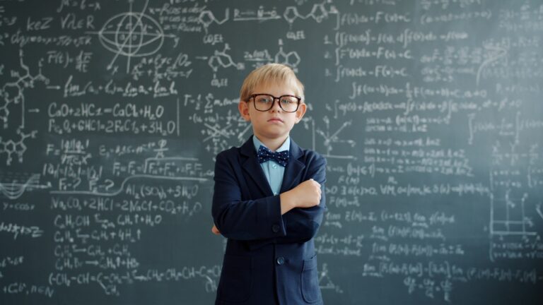 Young boy in glasses stands by chalkboard with equations
