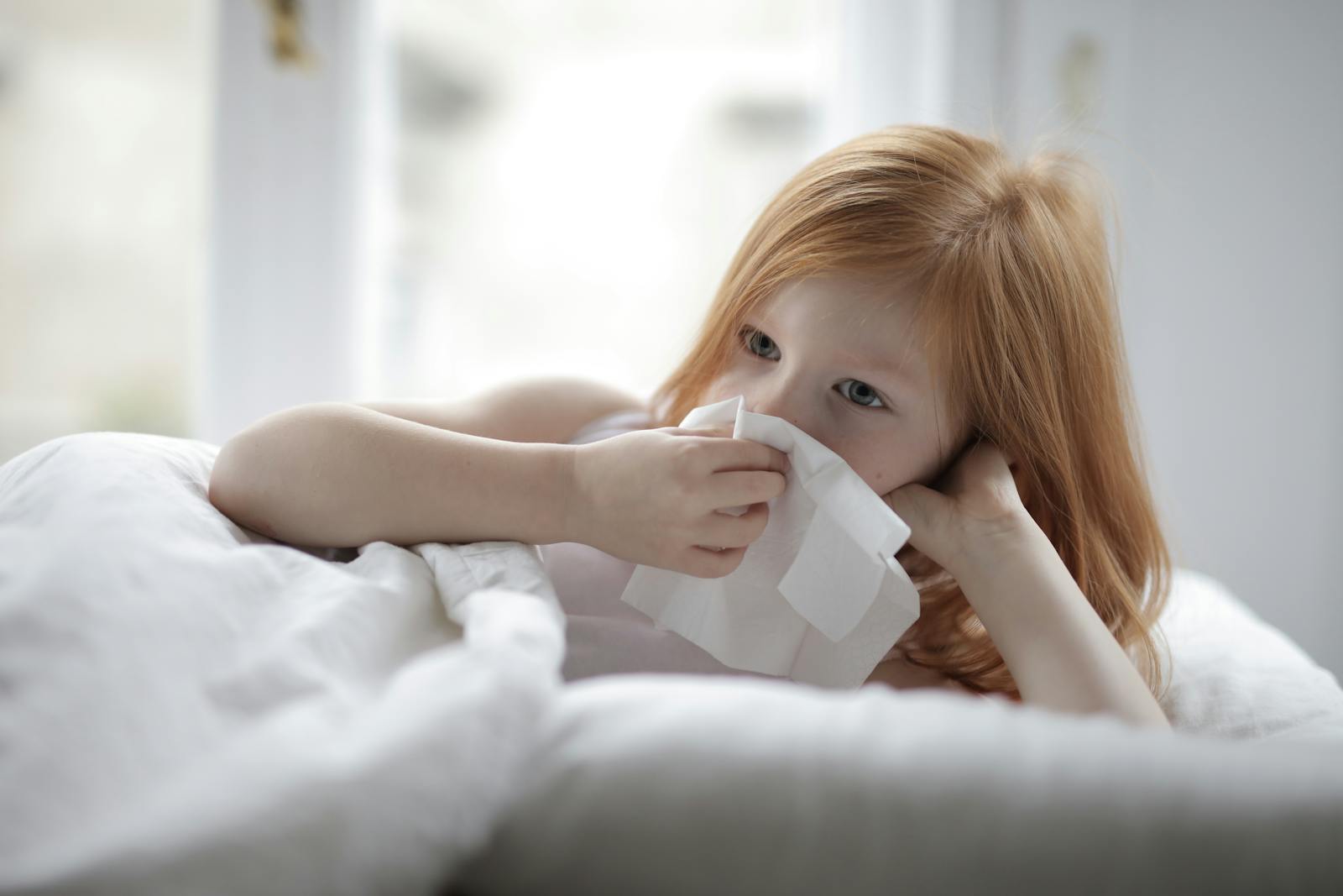 A young girl with red hair is lying in bed, using a tissue, suggesting she might be unwell.