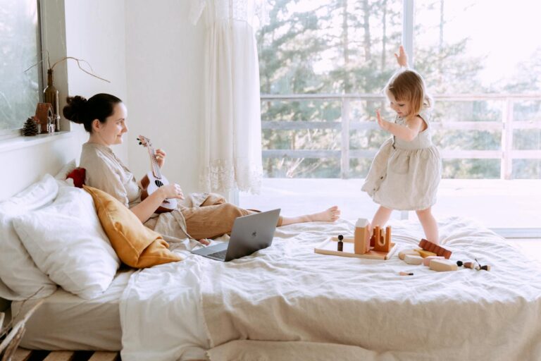 A joyful mother plays ukulele while her daughter dances in a sunlit bedroom.