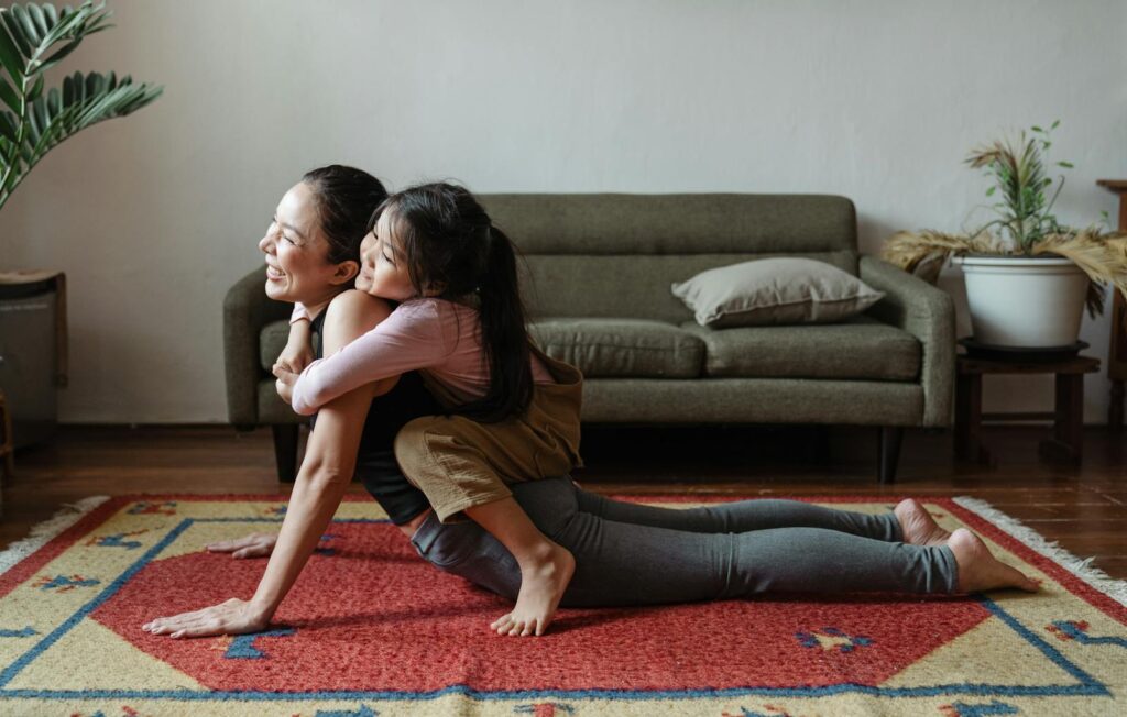 A woman practicing yoga with her daughter in a cozy living room, embracing and smiling.