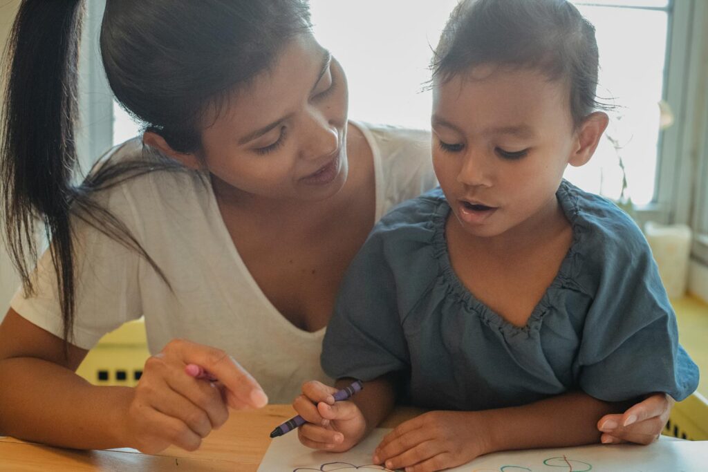 Content young ethnic female mother and cute daughter sitting at table and drawing on paper sheet with crayons while spending time together at home