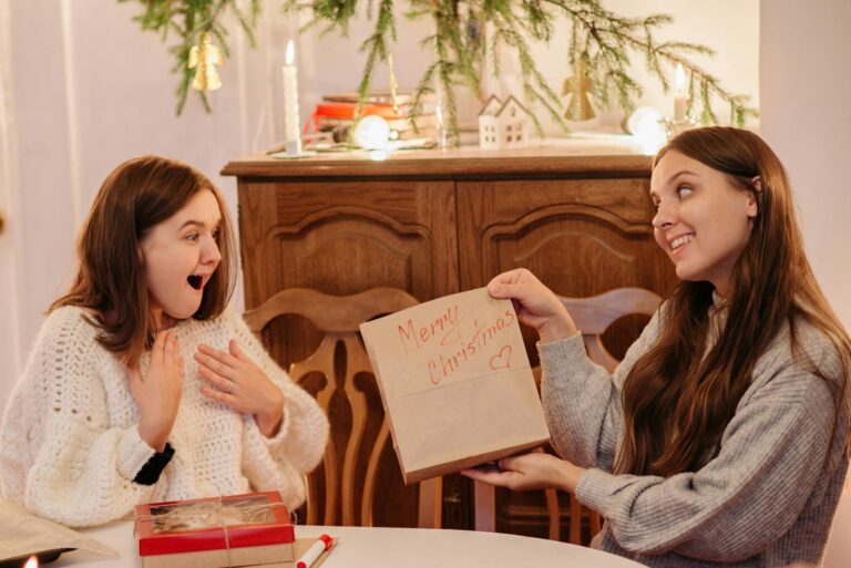 Two women exchanging Christmas gifts with joy and surprise indoors by a cozy setting.