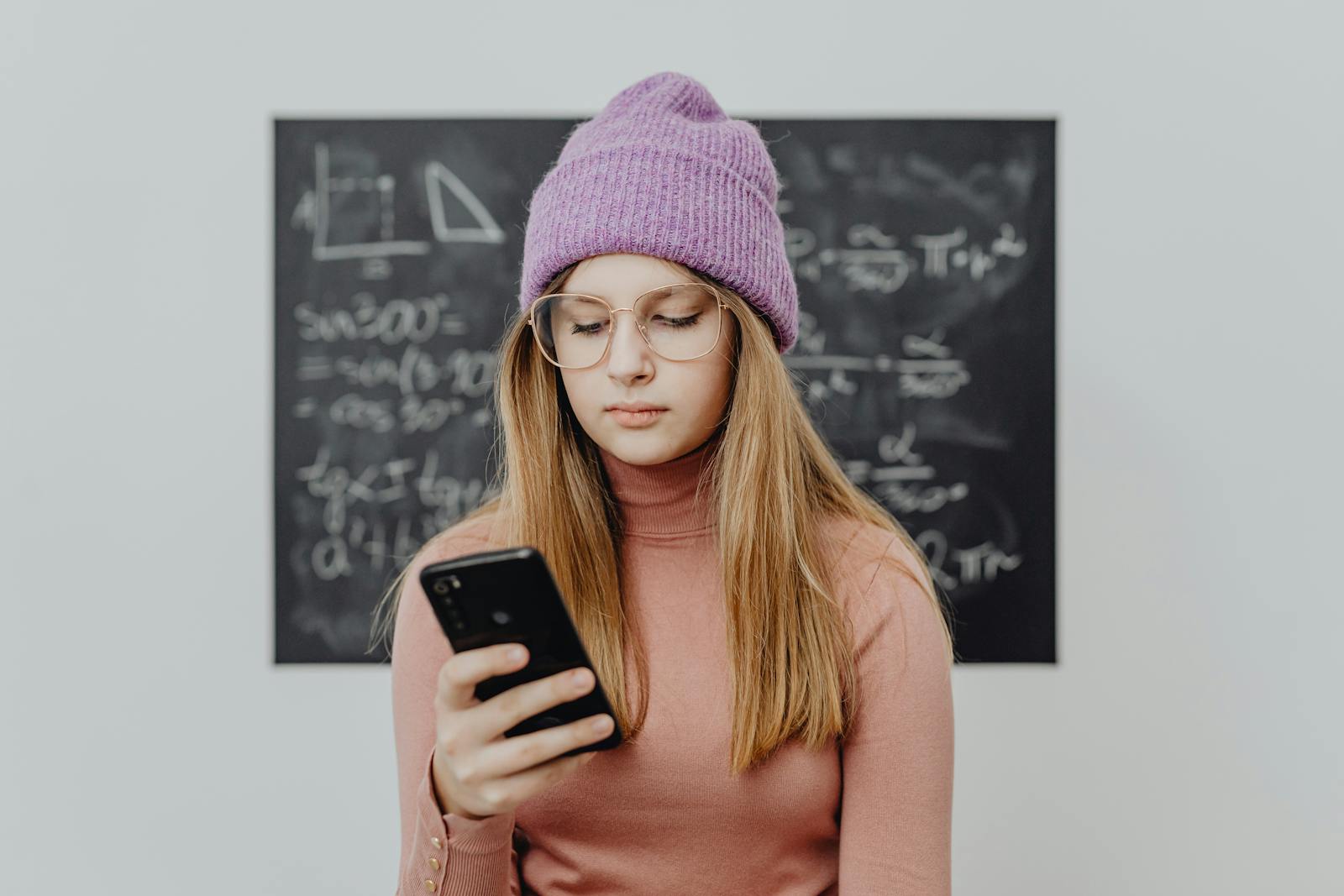 Teenager wearing a beanie and eyeglasses using a smartphone against a blackboard.