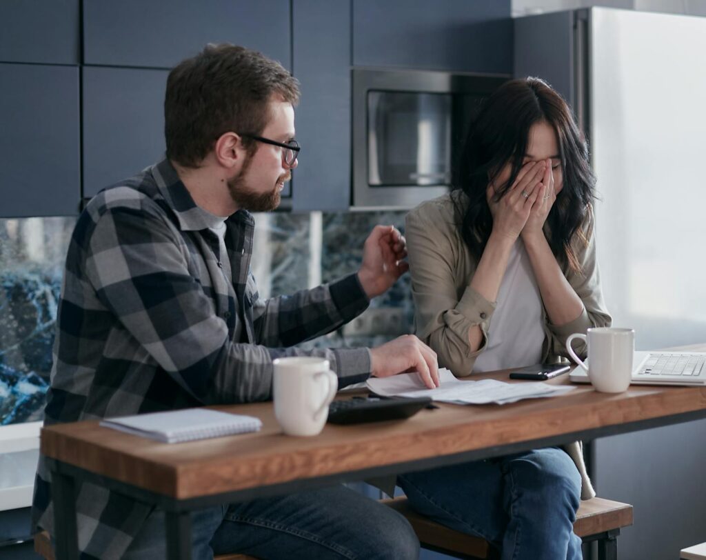 A couple discusses financial issues with bills and a laptop on the table, inside a modern kitchen setting.