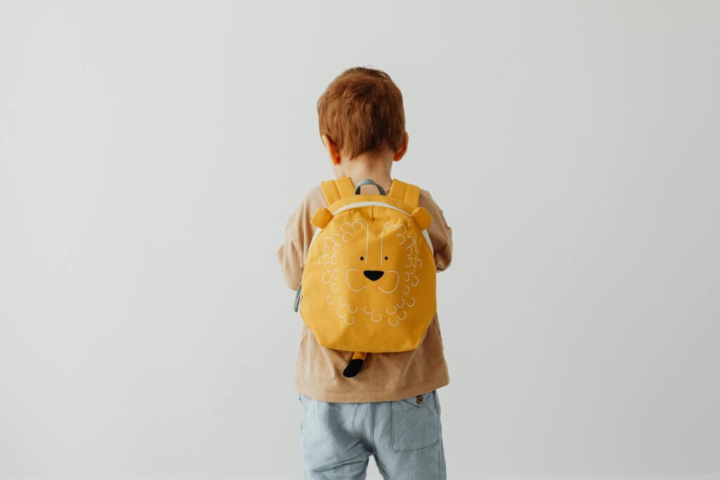 Cute child with red hair wearing a playful lion backpack facing away.