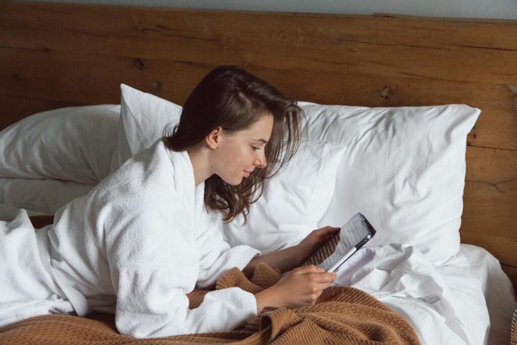 Woman in a robe reading a tablet in bed, surrounded by cozy pillows.