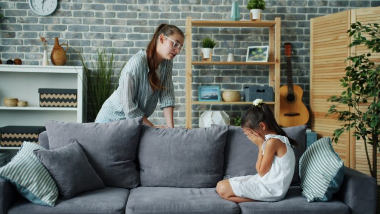 Mother scolds crying daughter on sofa in living room.