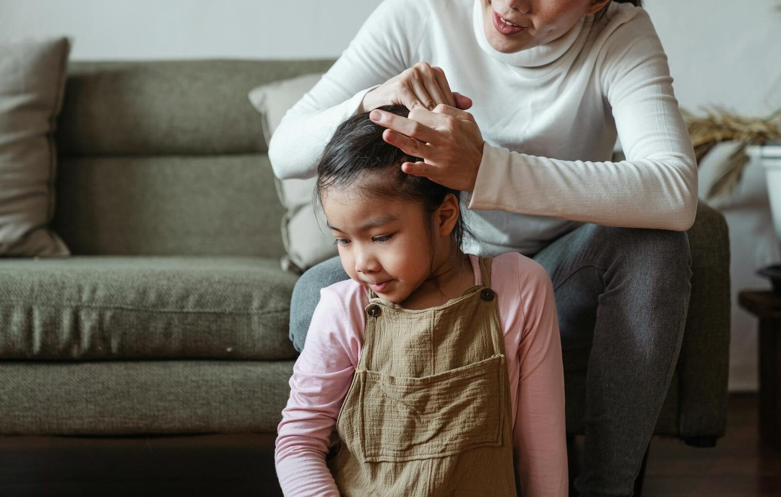 A mother lovingly ties her daughter's hair while sitting on the couch, sharing a tender bonding moment.