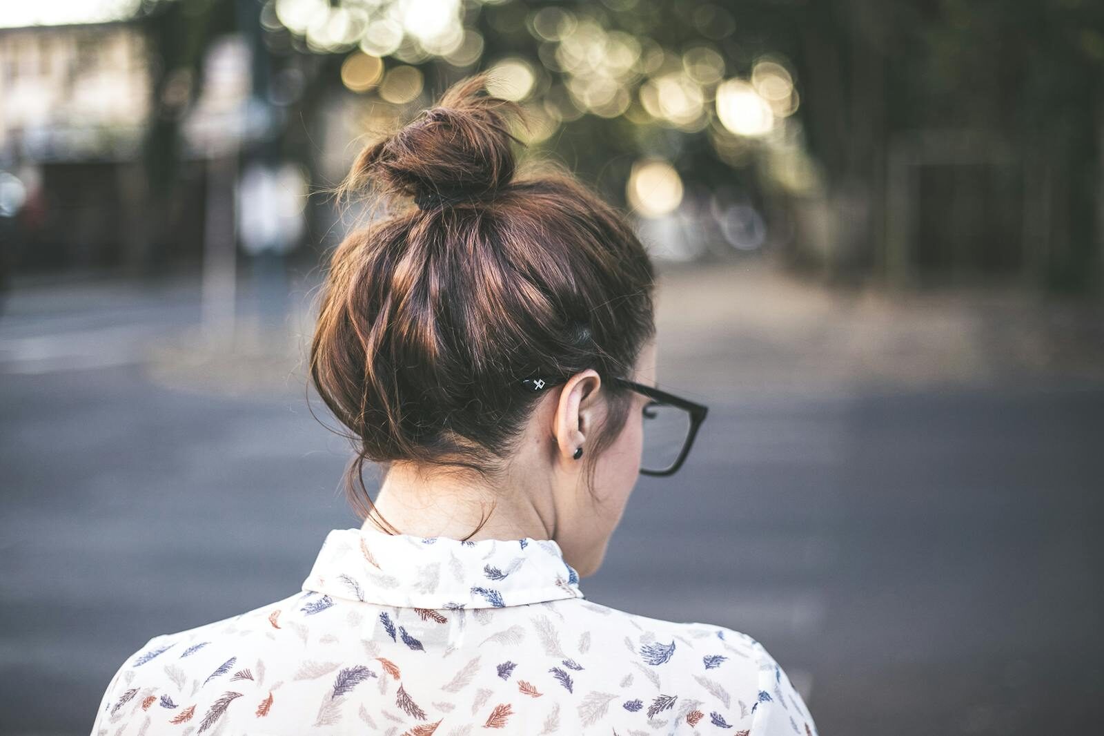Back view of a woman with glasses and a bun in a natural, blurry background.