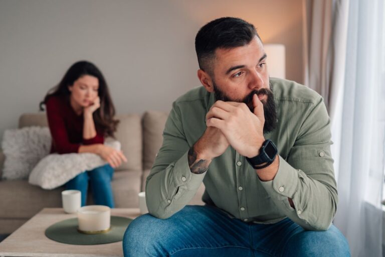 Man looking upset, woman behind him sitting on the couch