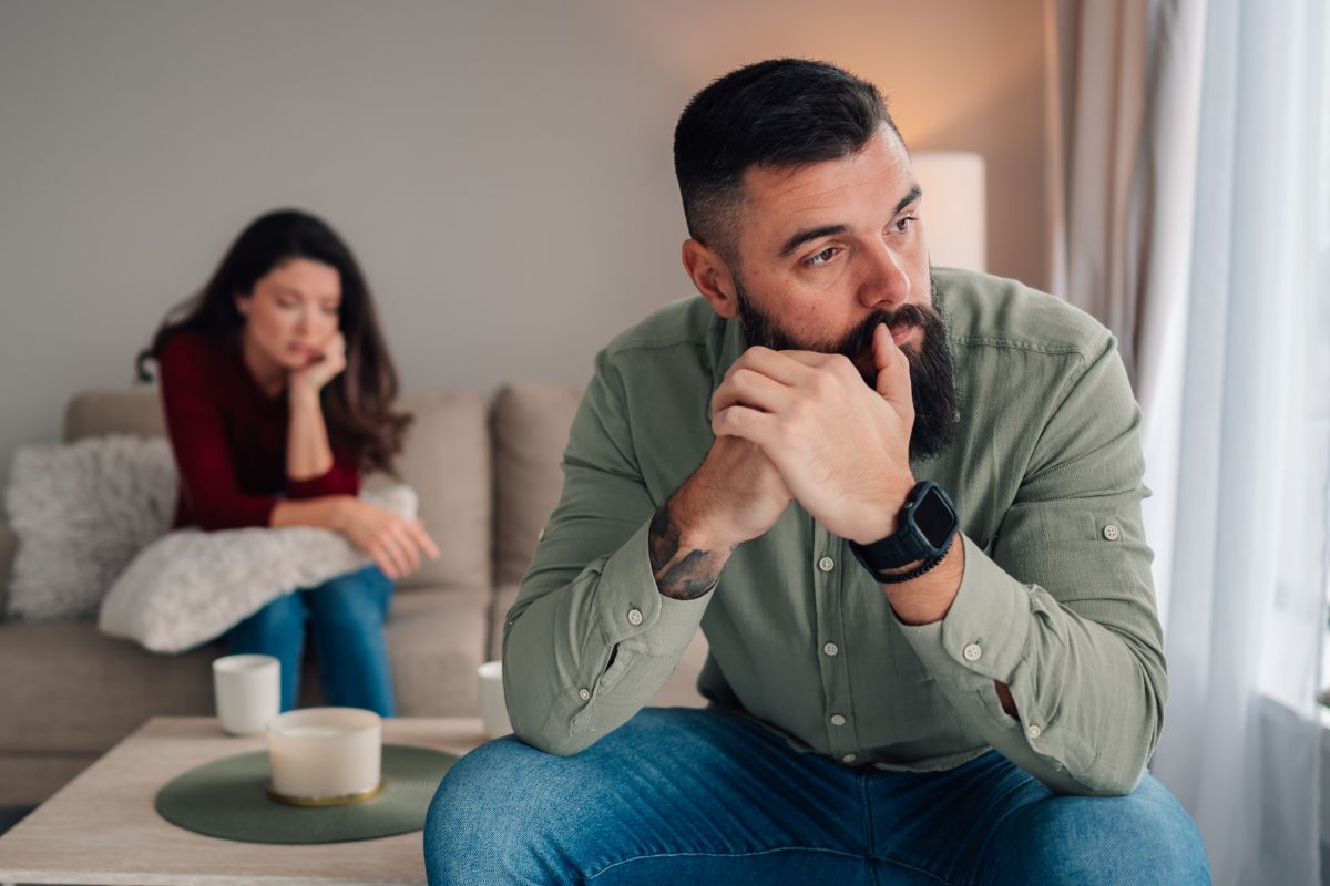Man looking upset, woman behind him sitting on the couch