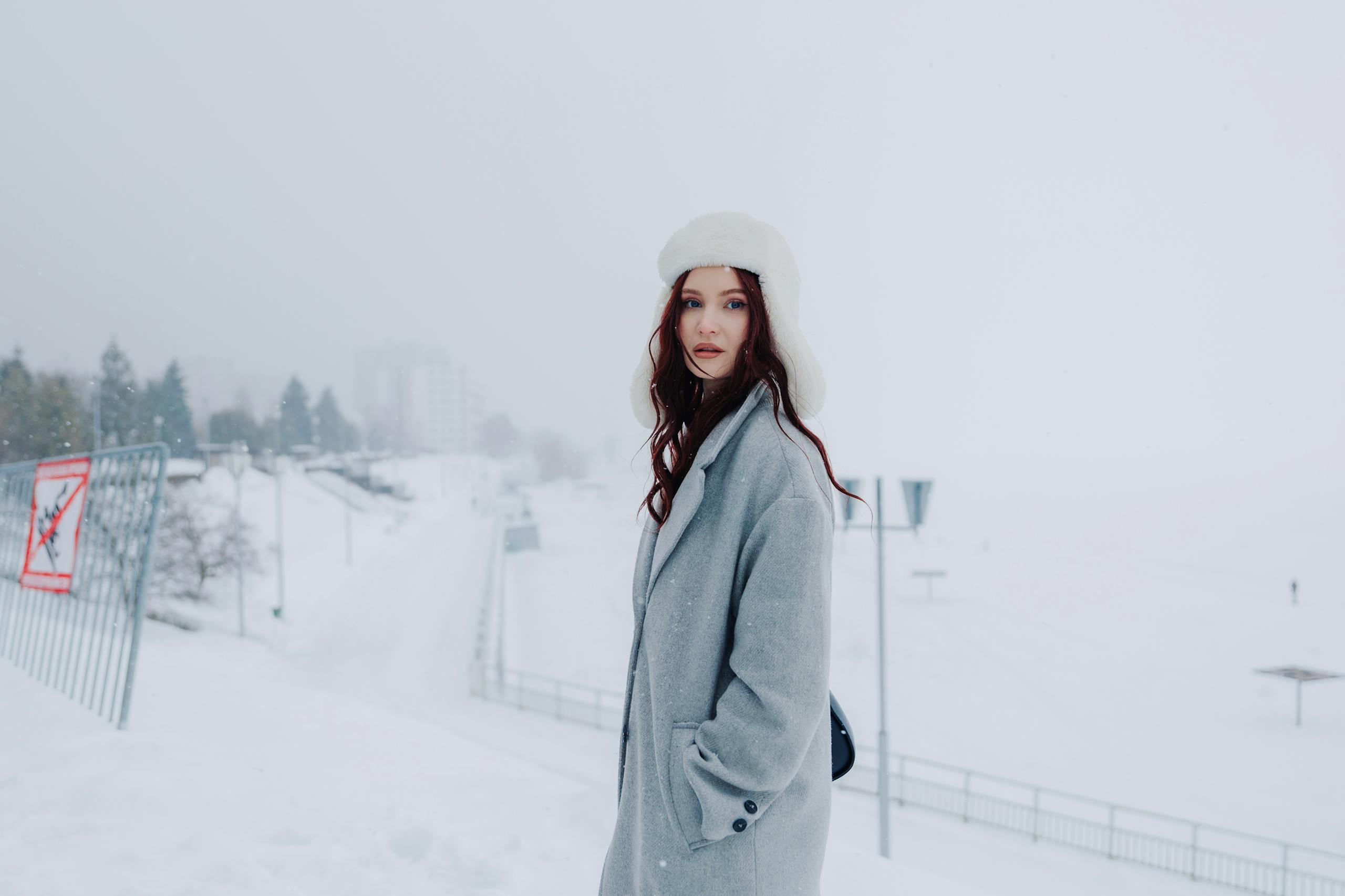 A fashionable woman wearing a grey coat and white hat standing in a snowy urban setting.