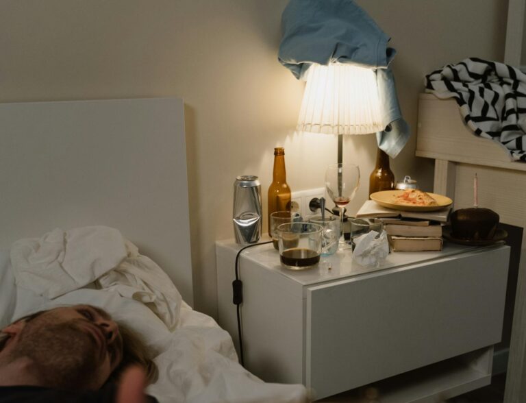 A man lies on a bed holding a guitar in a cluttered bedroom, suggesting a relaxed vibe.