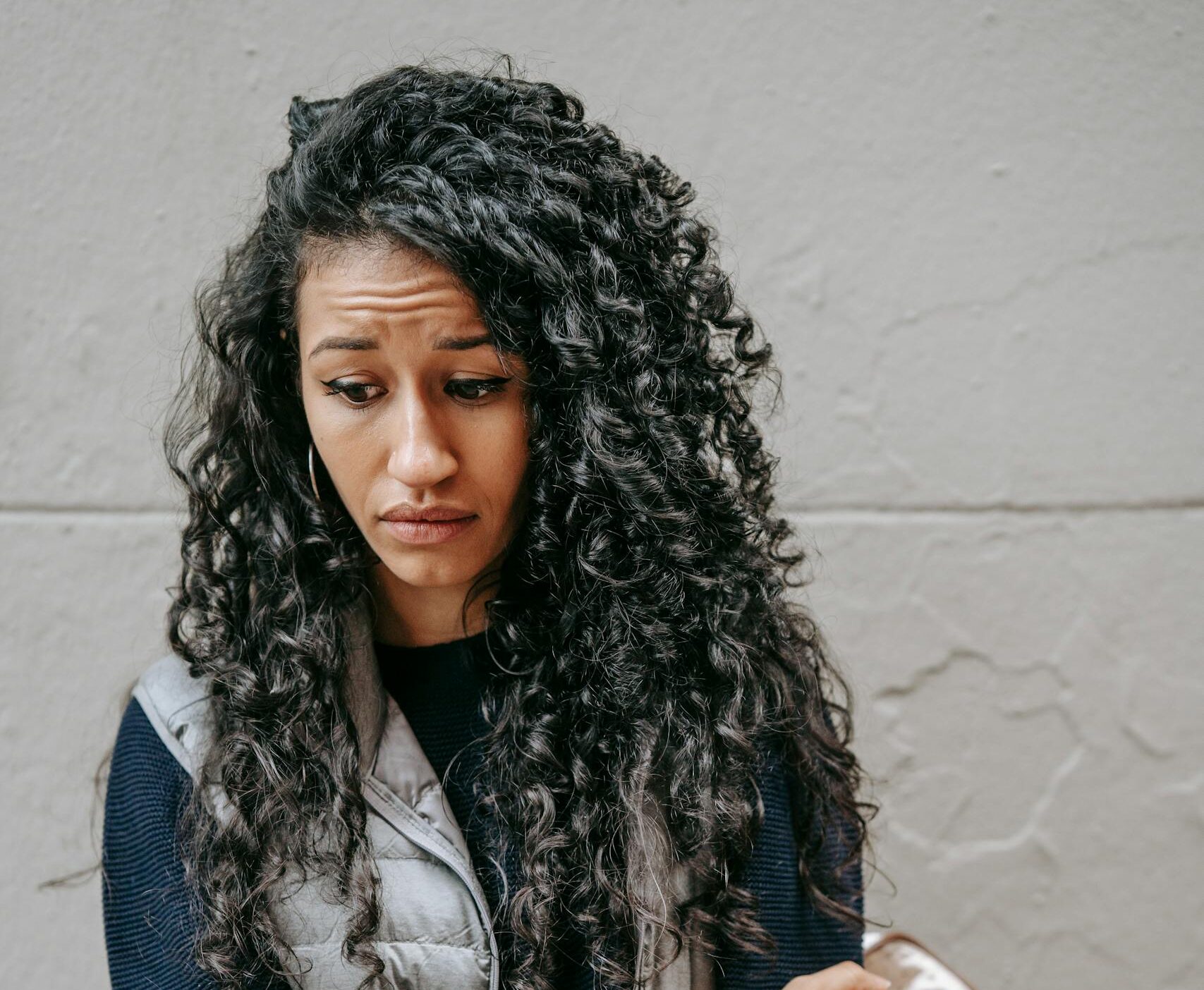 Upset young ethnic female with long curly hair in casual outfit standing on street and looking down thoughtfully