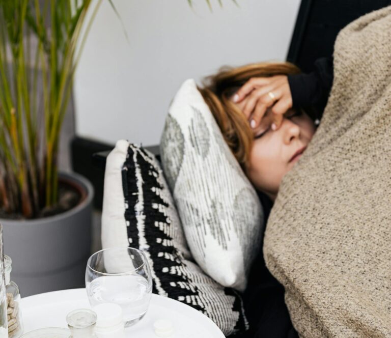 Woman lying on a couch covered with a blanket, feeling unwell, with pills on a table nearby.