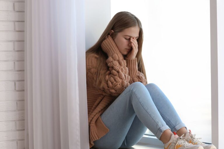 teen sitting on a window sill face in her hands sad