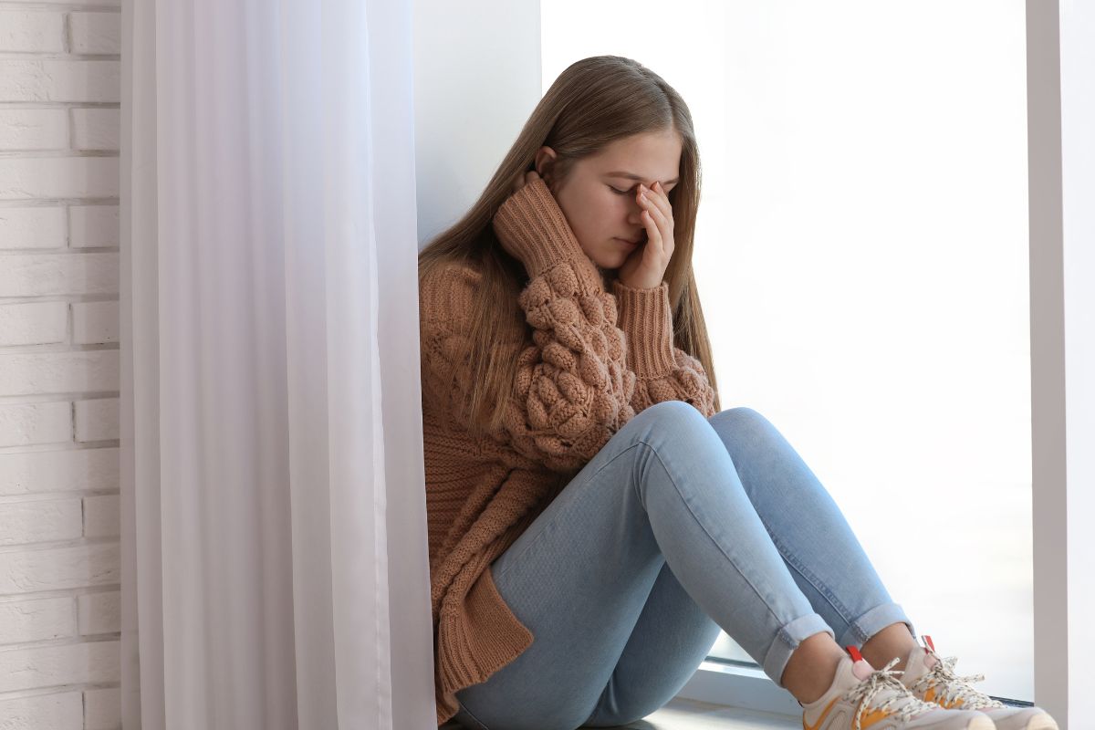 teen sitting on a window sill face in her hands sad